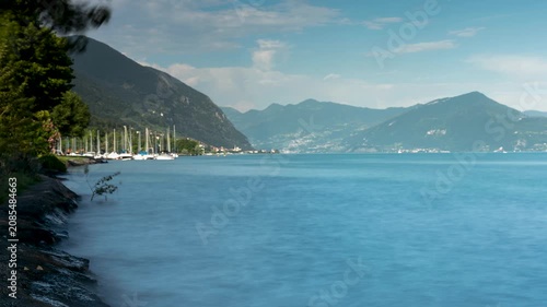 Beautiful time lapse of scenic Lake Iseo shore with moving clouds over green mountains, docked sailboats, and a calm turquoise water surface in Italy