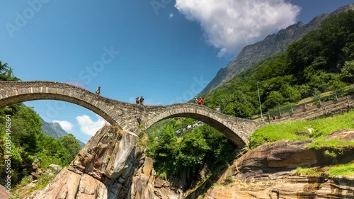 Timelapse of Ponte dei Salti double-arch stone bridge over emerald Verzasca river in Lavertezzo Switzerland with rushing clouds and tourists