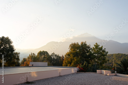 Soft sunrise light over a calm rooftop terrace, framed by trees and distant mountains. A serene, peaceful morning scene that evokes quiet reflection and outdoor relaxation.