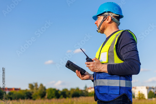 Tableau sur toile Engineer or utility worker wearing a blue hard hat and reflective safety vest, holding a digital tablet while holding a walkie-talkie transceiver outdoors