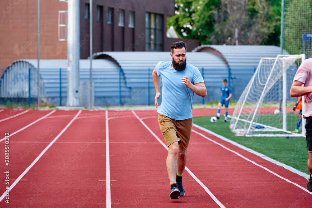 Obraz premium A man jogging on a red running track near a sports field, showcasing healthy exercise and outdoor activity with others playing in the background. A setting focused on activity and recreation.