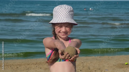 Smiling girl in panama hat holding sand ball on beach. Concept of kids summer seaside activities