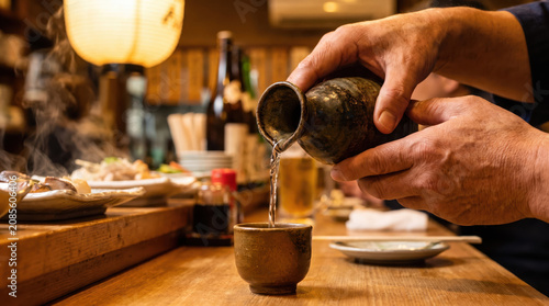 Sake hand cup pour bottle bar steam lantern wooden restaurant warm hand pour sake from ceramic bottle into small cup on wooden counter with steam and lantern in cozy restaurant