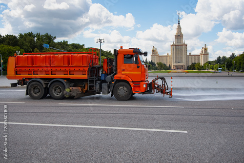 An orange municipal service truck sprays water onto a road for cleaning or dust control on the main street in front of Moscow University - Moscow, Russia