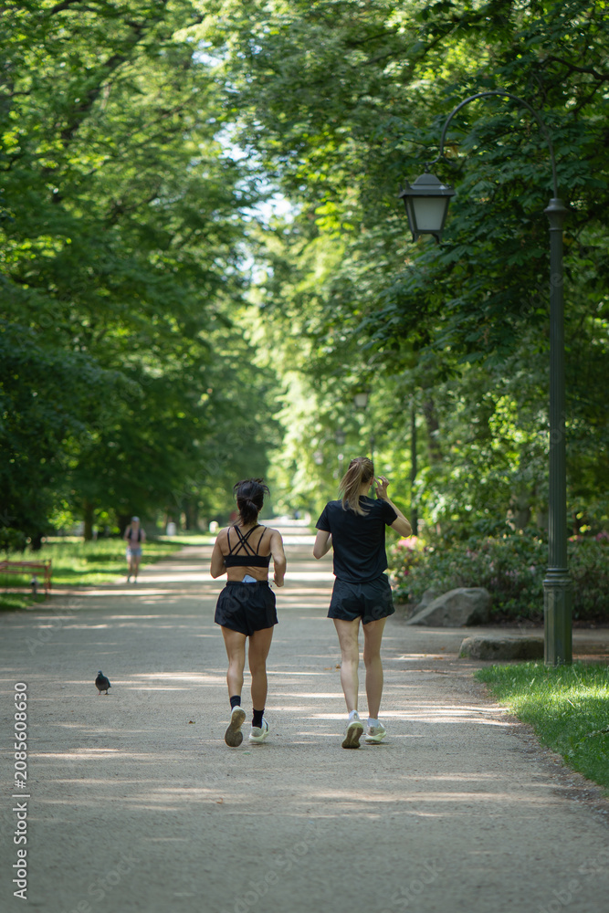 Fototapeta premium Two diverse fitness girlfriends running on path in public park. Active ladies training outdoor for healthy lifestyle. Group of women doing cardio workout exercise in nature on sunny summer day.