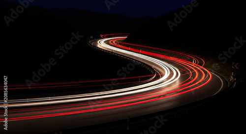 A long exposure photograph captures a winding road at night. The headlights of a vehicle streak across the frame, creating a vibrant, flowing trail of light.