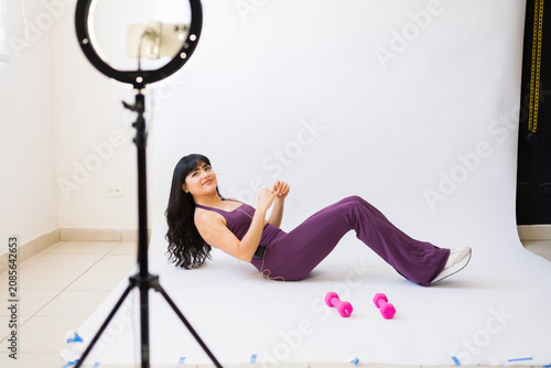 Fit woman in sportswear recording her virtual fitness class, performing exercises with dumbbells in a home studio setup