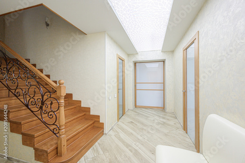 narrow hallway features a wooden staircase with ornate wrought‑iron railings, light wood flooring, textured off‑white walls, frosted‑glass doors, and a patterned ceiling panel