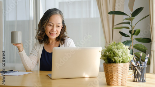 Canvastavla Mature woman relaxing with a cup of coffee while sitting at a home office desk with a laptop in a bright, cozy workspace