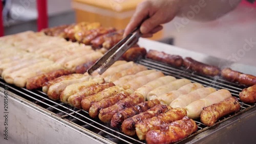 Various sausages are being grilled on a metal grill, with a hand using tongs to handle the food during cooking.