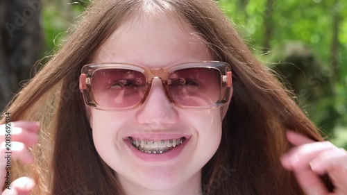 Young girl with dental braces put on sunglasses smiling looking at camera. Happy caucasian teenager outdoors on sunny summer day