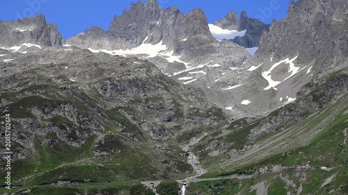 Scenic alpine valley with winding pass road and cascading waterfall amid green slopes and rocky peaks in summer.