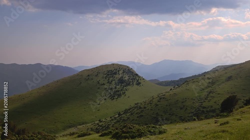 Time-lapse capturing soft evening sunrays filtering through overcast clouds, casting moving shadows and light accents across a beautiful green hill and layered distant mountain ridges during sunset.