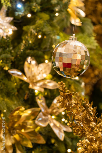 A close-up of a mirrored disco ball ornament hanging from a beautifully decorated Christmas tree, surrounded by golden floral decorations, warm fairy lights, and festive greenery