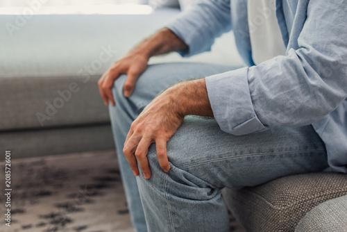 Foto A casual man sits on a sofa with hands resting on his knees, conveying a quiet moment of thought, waiting or relaxation in a comfortable home setting with denim jeans and a light shirt