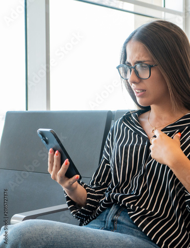 Bild auf Leinwand Young woman sitting in an airport lounge, looking at her smartphone screen with
