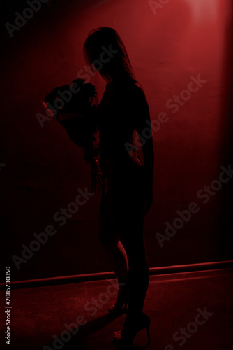 Full length silhouette portrait of a slender young woman holding a bouquet of roses against a black background with dramatic red illumination