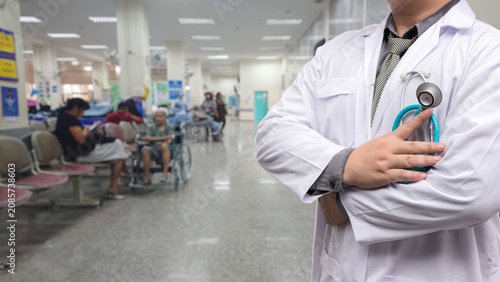 Close-up of male doctor in white coat holding a stethoscope with arms crossed. Blurred background of a busy hospital waiting room with patients sitting in chairs and wheelchairs.