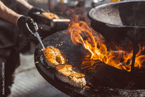 Salmon steaks grilling on large metal fire pit surface. Chef preparing fish steak over open flame. Gourmet street food cooking process in outdoor kitchen setting.