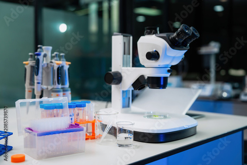 Compound microscope rests on a lab bench. Colorful plastic containers hold test tubes and beakers. Various lab equipment lines the workspace. Organized scientific tools feature in the modern lab