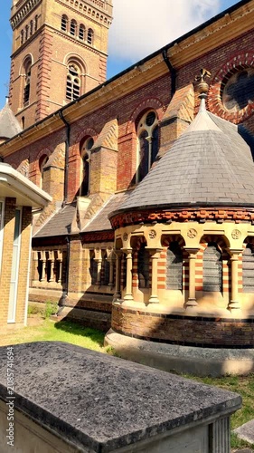 Upward tilt shot of the tower and rear exterior section of a brick built, ornate, 19th century, historic English church, bathed in sunshine, against a blue sky.