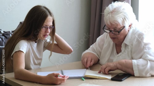 Senior woman and young girl wearing glasses sitting at table doing homework together. Grandmother explaining schoolgirl home tasks