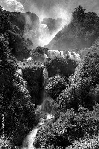 Vertical black and white view of Marmore Falls in Umbria, Italy. Multiple waterfalls cascade through dense forest while mist rises from the powerful water, creating a dramatic and timeless natural lan