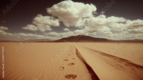 Footprints lead across vast desert sand toward distant mountain under dramatic cloud filled sky, evoking solitude and quiet wanderlust