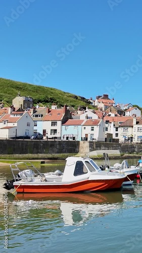 Panning shot across Staithes waterfront, harbour and fishing boats on the North Yorkshire coast, showcasing colourful cottages, sparkling water, and bright sunshine on a beautiful summer day.