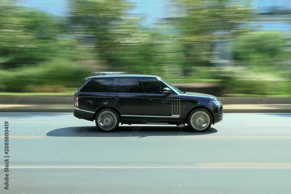 Fototapeta premium Black SUV car driving fast on suburban asphalt roadside with green trees
