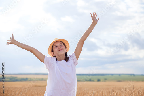 Young girl wearing a straw hat raises her arms joyfully in a golden wheat field under a cloudy sky, with green hills visible in the background