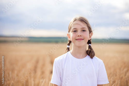 Young girl with pigtails wearing a white t-shirt stands in a golden wheat field under a cloudy sky, smiling softly at the camera with a serene landscape behind her