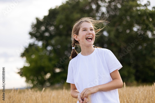Young girl with pigtails in a white t-shirt stands in a wheat field, smiling against a backdrop of trees and a cloudy sky