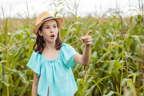 Young girl in a straw hat gestures while standing amidst tall corn plants in a field, showcasing her enthusiasm and the lush greenery around her
