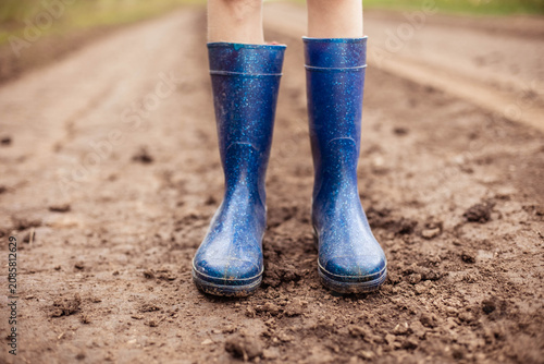 Blue rubber rain boots worn by a person standing on a muddy dirt path with greenery visible in the background, showcasing outdoor footwear suitable for wet conditions