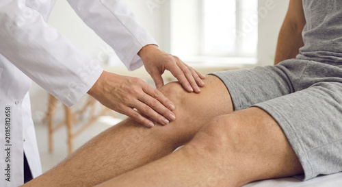 Doctor wearing white lab coat examining male patient knee in clinical medical therapy office room set.