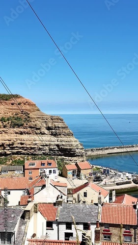 Vertical panning shot from a high viewpoint over Staithes fishing village on the North Yorkshire coast, showing rooftops, cliffs, and sea under bright summer sunshine.