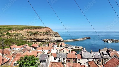 High-angle panning shot over Staithes fishing village on the North Yorkshire coast, showing rooftops, cliffs, and sea under bright sunny summer skies.