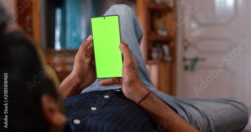 Obraz na plátně Man Lying Down at Home Holding Smartphone with Blank Green Screen Chroma Key Mockup for Apps Display