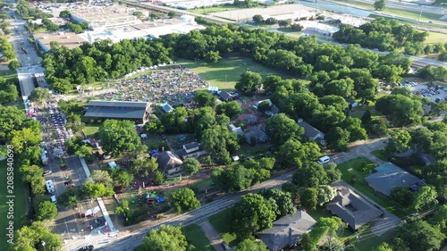 July Fourth Independence Day visitors populate shaded grounds within Farmers Branch Historical Park. Lush dense forest foliage borders organized event spaces across suburbs Dallas Fort Worth, TX