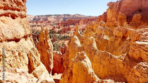 A smooth cinematic low angle shot panning across unique red rock spires resembling chess pieces or statues in Bryce Canyon, Utah. Scenic hiking trails and rare pine trees under a clear blue sky.