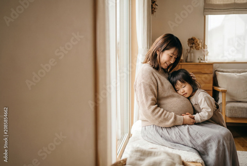 Pregnant woman sitting indoors with child embracing her belly  