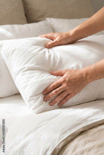 Hands fluffing a soft white pillow on a neatly made bed  