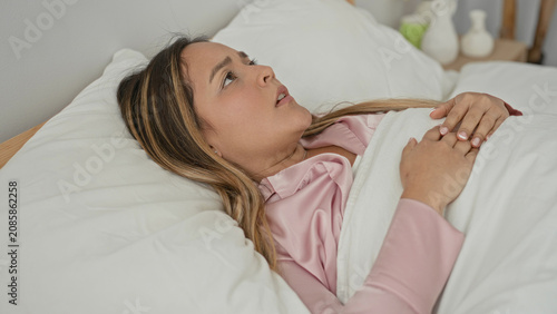 Foto Young hispanic woman in pink pyjamas in bed clutching a white pillow and pulling the duvet, grimacing with hand on head beside wooden frame in a building; restless insomnia