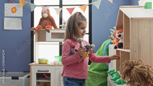 Konstfotografi Girl holding several dolls and reaching for a small toy on a wooden shelf in a colorful playroom inside a building; playful curiosity
