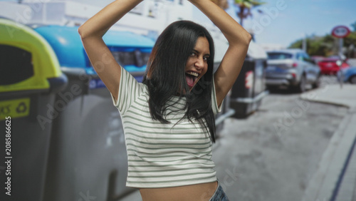 Fotografie Woman with arms raised above head beside large recycling bin, mouth open and tongue out in a spirited pose on a crowded urban street; playful joy