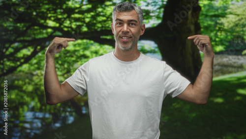 Bild auf Leinwand Man in white tshirt points fingers to chest with smirk, bare forearms visible, standing by large tree and pond in forest; confidence self
