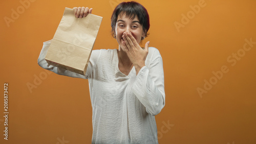 Valokuva Woman holding paper bag aloft with hand covering mouth, smiling and surprised in a studio orange backdrop mid reveal; surprise