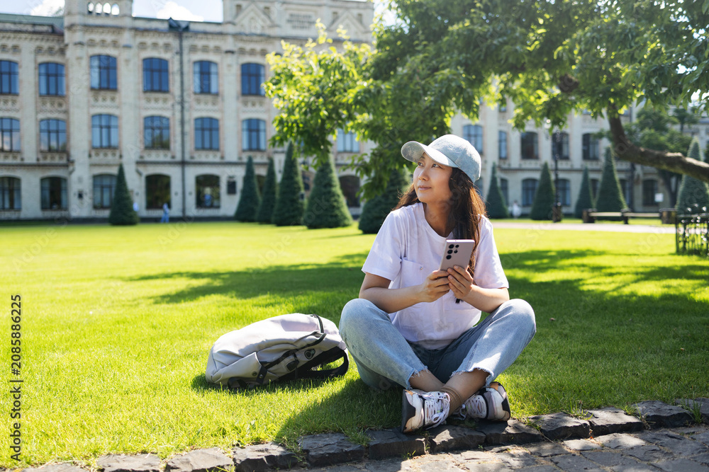 Fototapeta premium Young asian student woman smiling happy using smartphone sitting on the grass at the university campus