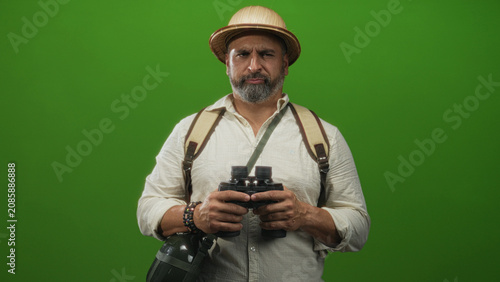 Canvastavla Man with grey beard holding binoculars and backpack, wearing pith helmet and bracelets against a vivid green studio wall; curiosity travel exploration